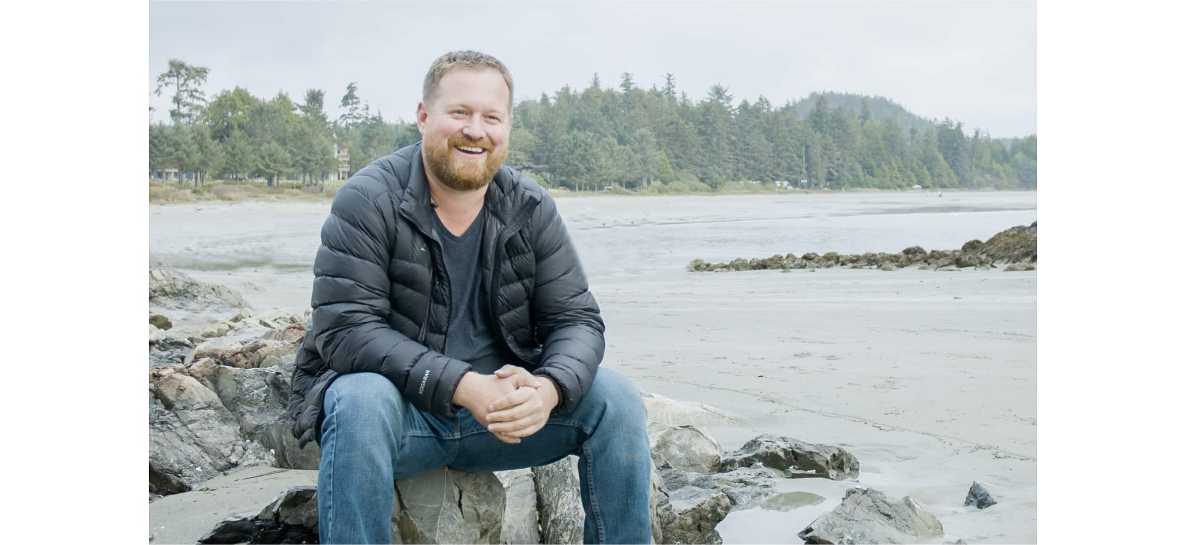 Chad sits happily on some rocks by a river lined with trees.